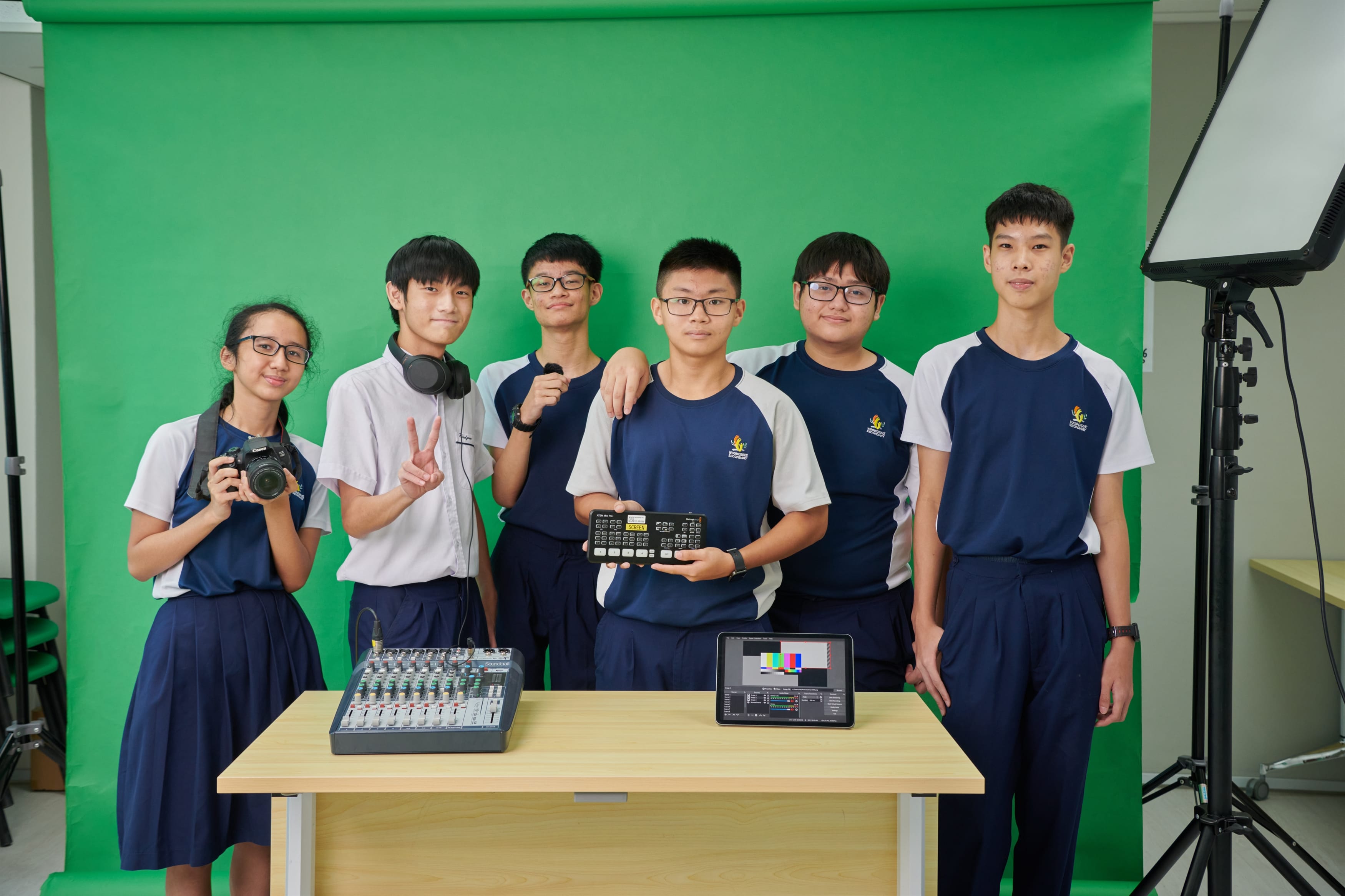 Students pose before a green screen with a camera, audio mixer, tablet, and studio light.